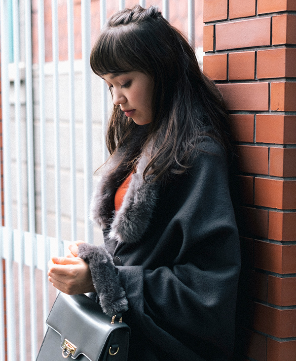 レンガの壁に軽く体を預け誰かを待っている女性の写真